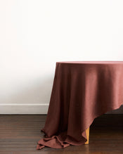 A wooden table, partially covered by the Bed Threads Cacao & Hazelnut 100% French Flax Linen Table Bundle, sits on a dark wooden floor against a plain white wall. - view