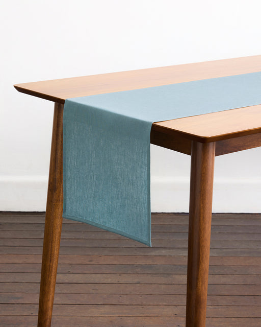 A Lagoon 100% French Flax Linen Table Runner by Bed Threads in light blue is neatly laid along the center of a rectangular wooden table with angled legs, set on a dark wood floor against a white wall.