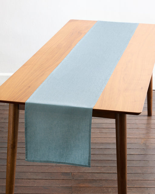A wooden table with angled legs is topped with a Lagoon 100% French Flax Linen Table Runner by Bed Threads, draping over one end. Set on a wooden floor against a white wall, it brings simple elegance to your dining space.