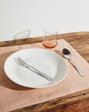 The Hazelnut & Terracotta 100% French Flax Linen Table Bundle by Bed Threads features a white plate with a fork and spoon, a clear glass cup, and classic placemats arranged on a rustic wooden table. - view