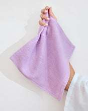 A modern white bathroom sink with a stack of Bed Threads’ Violet 100% Cotton Terry Face Washers (Set of Two), plus a beige soap bar, wooden brush, and a matching violet bath mat on the floor. - view