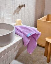 The Violet 100% Cotton Terry Hand Towel by Bed Threads sits on a white tiled sink beside a soap bar and glass of water, with a wooden stool and tub in the softly lit, quick-drying bathroom. - view
