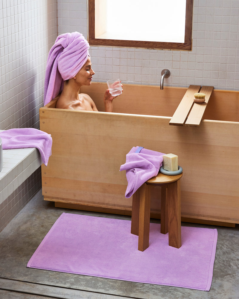 A person relaxes in a wooden bathtub, wearing a purple towel turban and holding a glass of water. Nearby, the Bed Threads Violet 100% Cotton Terry Bath Sheet is draped over a stool, with a matching bath mat on the floor.