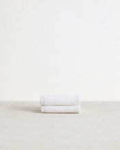 A set of two Crisp White 100% Cotton Terry Face Washers by Bed Threads, neatly folded with beige branded fabric labels in one corner, rests atop a light textured surface. - view