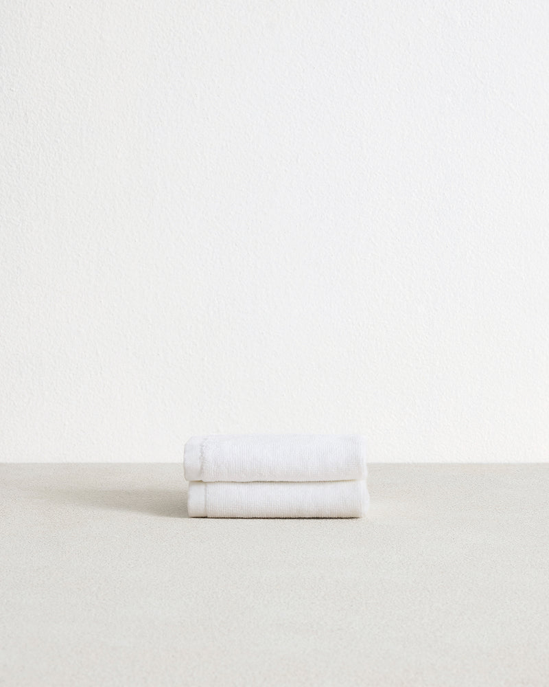 A set of two Crisp White 100% Cotton Terry Face Washers by Bed Threads, neatly folded with beige branded fabric labels in one corner, rests atop a light textured surface.