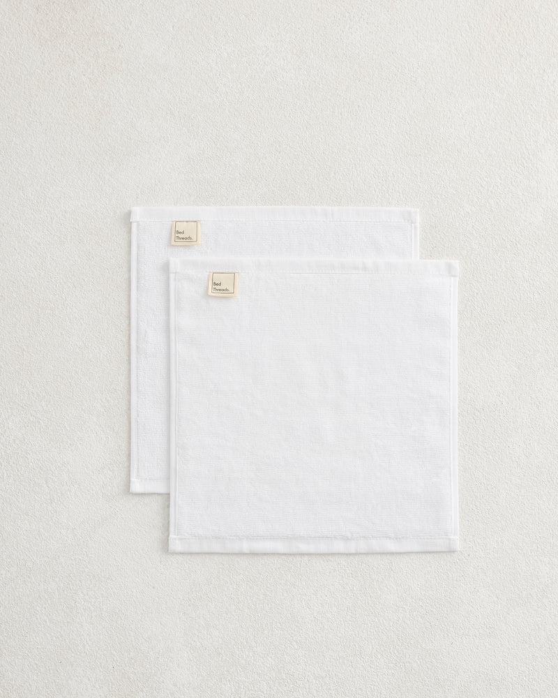 A set of two Crisp White 100% Cotton Terry Face Washers by Bed Threads, neatly folded with beige branded fabric labels in one corner, rests atop a light textured surface.