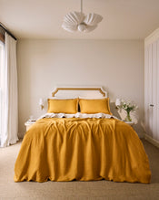 A neatly made bed with a beige and gold headboard, white sheets, a mustard yellow blanket, and Bed Threads Turmeric & Crème 100% French Flax Linen Swirl Pillowcases (Set of Two) in mustard yellow. - view