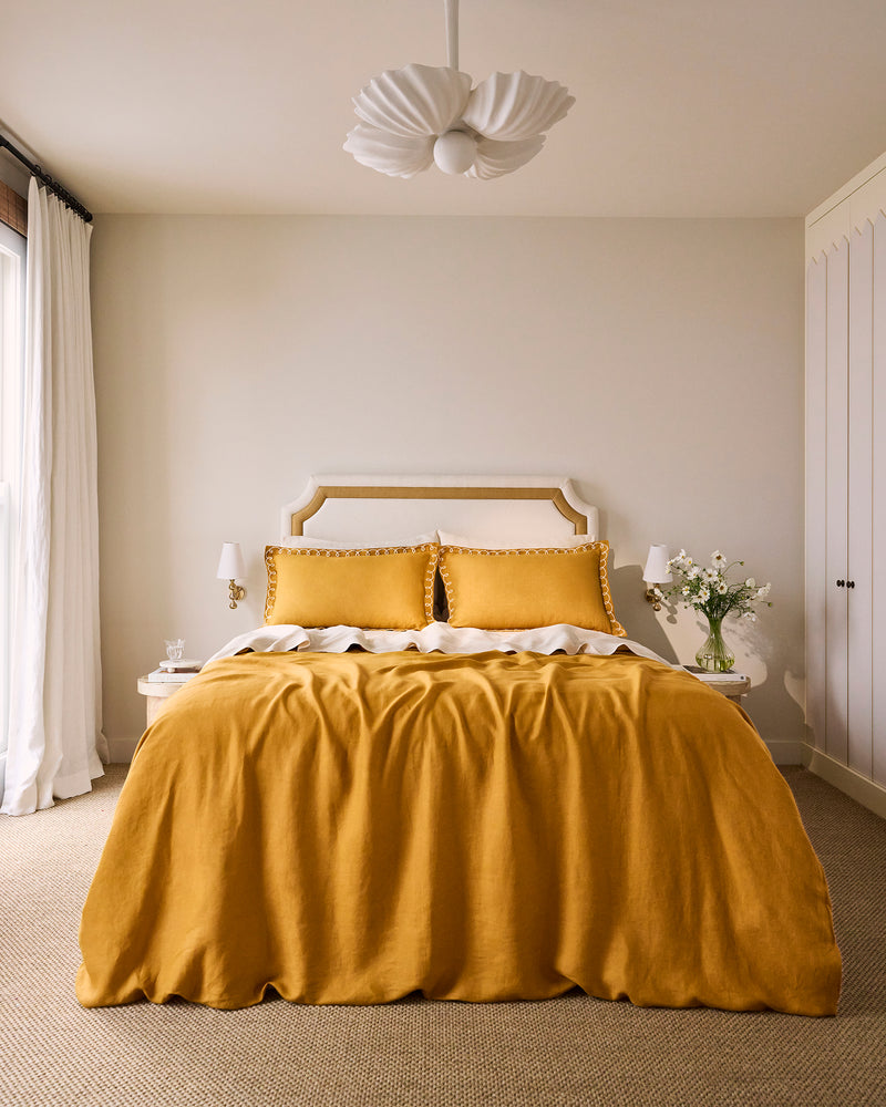A neatly made bed with a beige and gold headboard, white sheets, a mustard yellow blanket, and Bed Threads Turmeric & Crème 100% French Flax Linen Swirl Pillowcases (Set of Two) in mustard yellow.