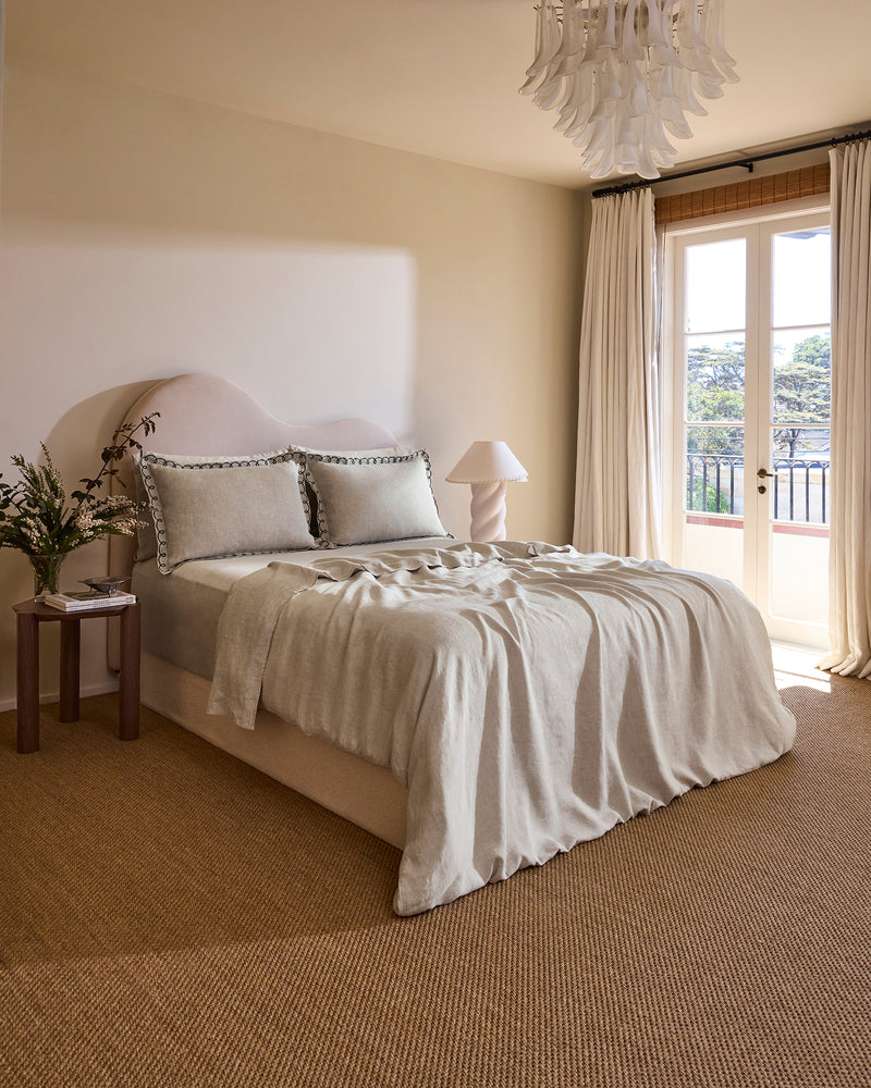 A neatly made bed with a beige upholstered headboard, two Bed Threads Oatmeal & Charcoal 100% French Flax Linen Swirl Pillowcases featuring black trim, and a matching slightly wrinkled French flax linen blanket.