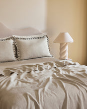 A neatly made bed with Bed Threads’ Oatmeal & Charcoal 100% French Flax Linen Swirl Pillowcases, set of two, sits in neutral tones beside a bedside table featuring a sculptural ceramic lamp and pleated lampshade. - view