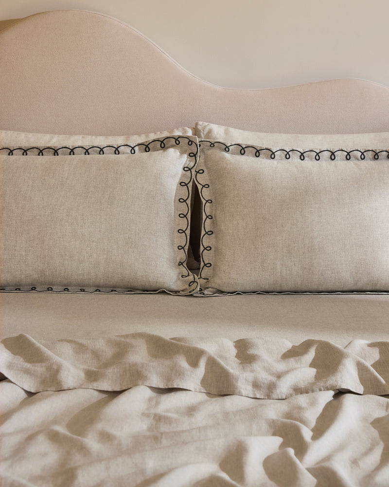 A neatly made bed with a beige upholstered headboard, two Bed Threads Oatmeal & Charcoal 100% French Flax Linen Swirl Pillowcases featuring black trim, and a matching slightly wrinkled French flax linen blanket.