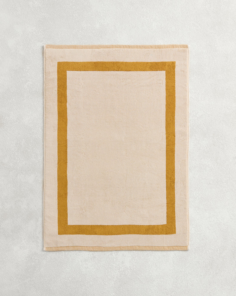 The Bed Threads Crème & Turmeric Two-Toned 100% Cotton Terry Bath Mat features a plush beige center with a bold mustard border, adding cozy style to your space.
