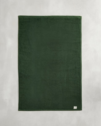 The Bed Threads Moss 100% Cotton Terry Bath Mat, in dark green, lies flat on a light gray surface with a small white tag visible in one corner.