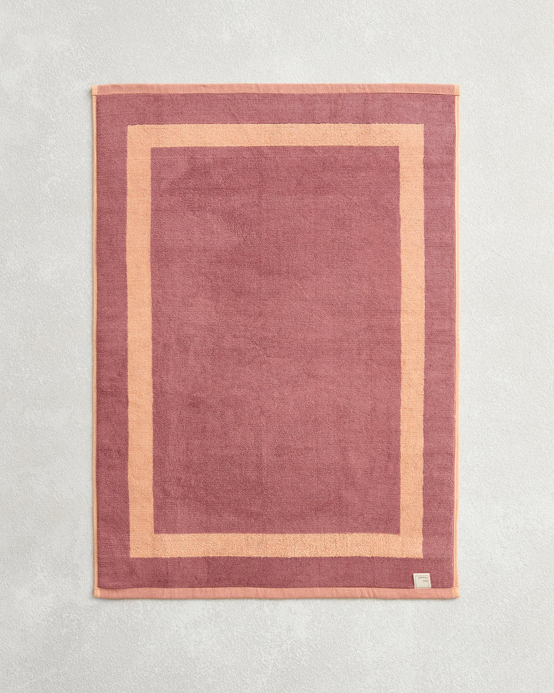 The Bed Threads Terracotta & Pink Clay Two-Toned 100% Cotton Terry Bath Mat, shown flat on a textured surface with a visible corner tag, pairs perfectly with plush towels for a coordinated bathroom style.