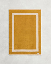 The Crème & Turmeric Two-Toned 100% Cotton Terry Bath Mat by Bed Threads, featuring a mustard yellow hue and simple white border, is shown on a light textured surface. - view