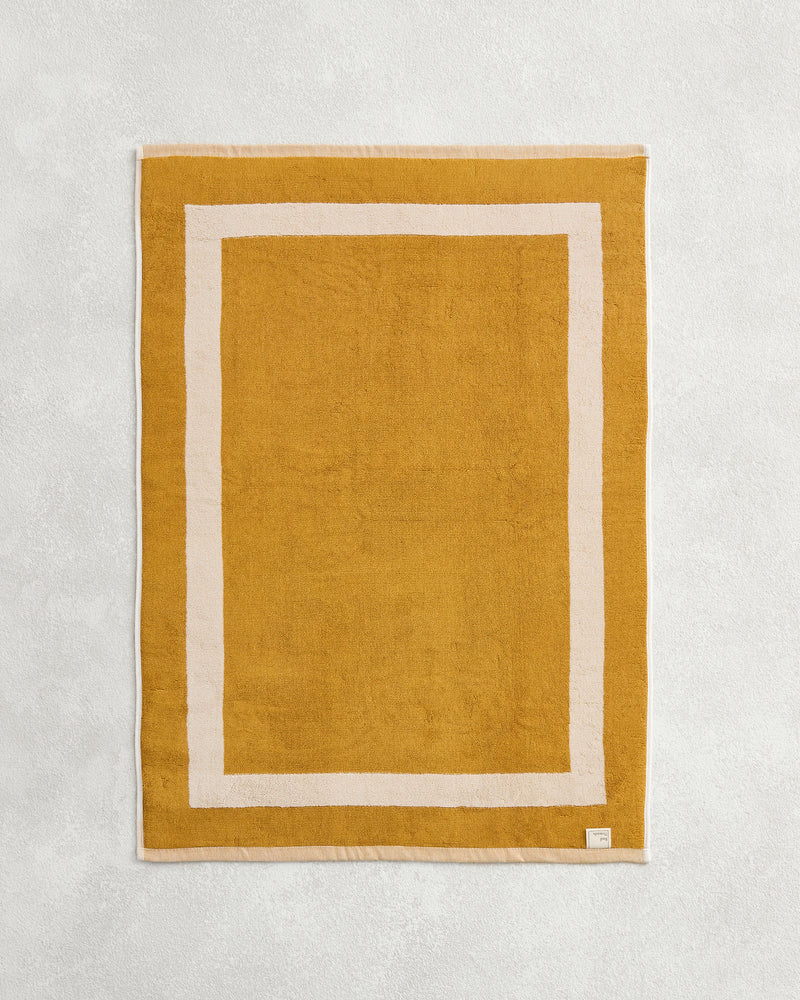 The Crème & Turmeric Two-Toned 100% Cotton Terry Bath Mat by Bed Threads, featuring a mustard yellow hue and simple white border, is shown on a light textured surface.