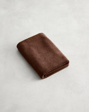 A neatly folded brown towel rests on a light gray, subtly textured surface next to the Bed Threads Espresso 100% Cotton Terry Bath Mat. - view