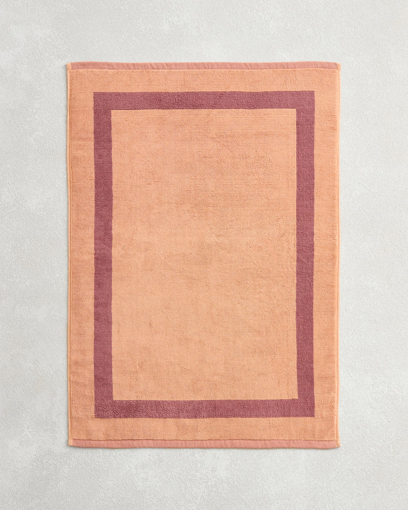 The Bed Threads Terracotta & Pink Clay Two-Toned 100% Cotton Terry Bath Mat, laid flat on a light gray textured surface, offers plush softness and a cozy feel with its warm hues.