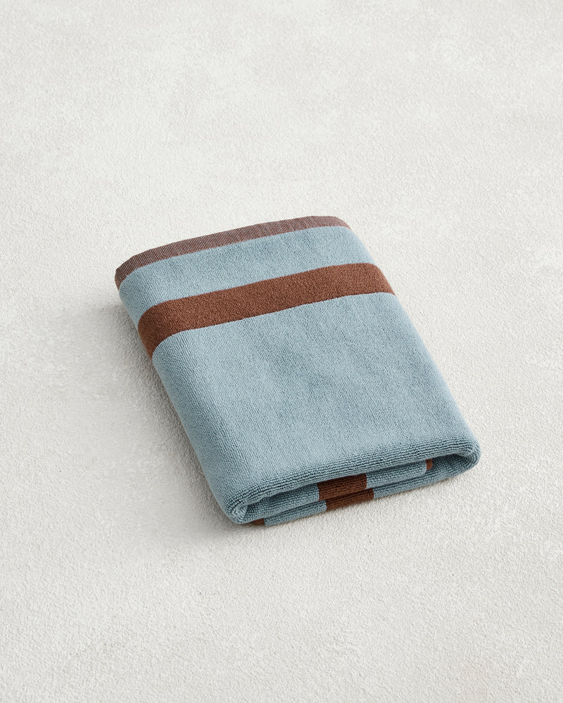 The Cacao & Lagoon Two-Toned 100% Cotton Terry Bath Mat by Bed Threads features a folded blue design with two horizontal brown stripes, shown on a light textured surface.