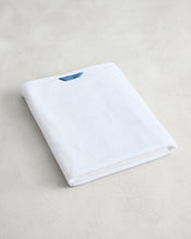 The Bed Threads Crisp White 100% Cotton Terry Bath Sheet, featuring a small blue tag, is neatly folded and displayed on a light-colored surface. - view