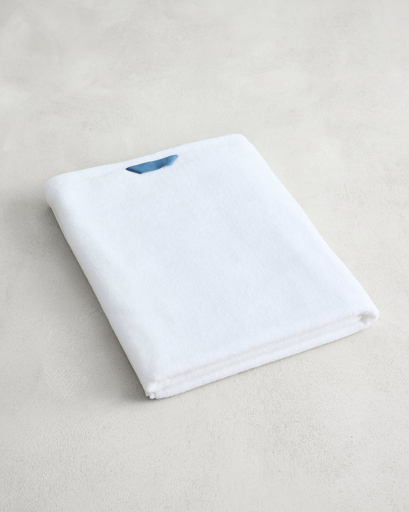 The Bed Threads Crisp White 100% Cotton Terry Bath Sheet, featuring a small blue tag, is neatly folded and displayed on a light-colored surface.