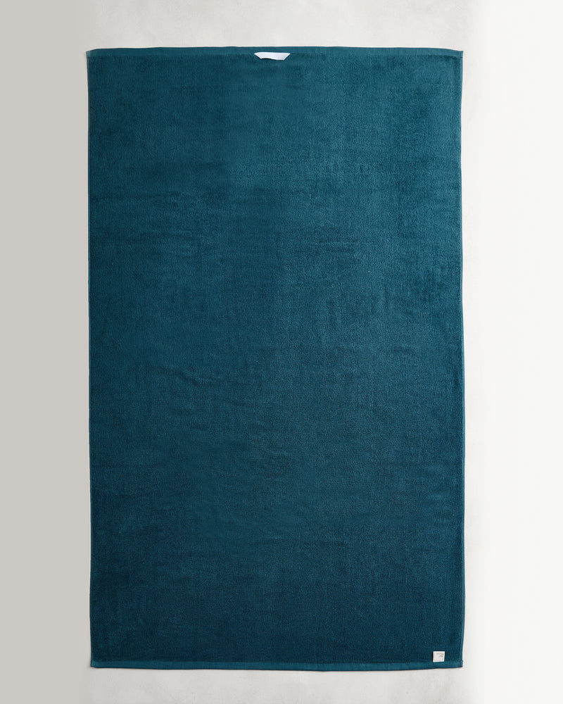 A large, rectangular dark teal Aegean 100% Cotton Terry Bath Sheet by Bed Threads lies flat on a light surface, featuring a white label at the top edge and a sewn-in tag at the bottom—perfect for lovers of luxe cotton towels.