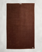 The Bed Threads Espresso 100% Cotton Terry Bath Sheet, large and rectangular with a plush texture, is laid flat on a light gray surface, showing a small white tag in one corner and a hanging loop at the top. - view