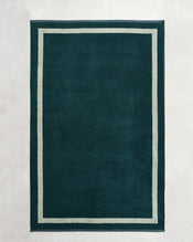 A dark green rectangular rug with a light border, inspired by the elegant Forest & Sage Two-Toned 100% Cotton Terry Bath Sheet Bundle from Bed Threads, lies on a pale, textured surface. - view