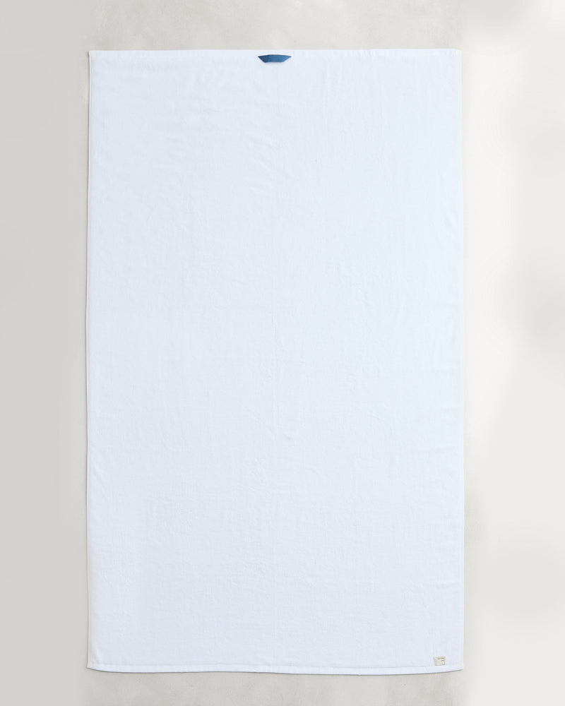 Two Crisp White 100% Cotton Terry Bath Sheets from Bed Threads are laid flat on a light surface, each featuring a small blue loop at the top edge and a discreet tag at the bottom right corner.