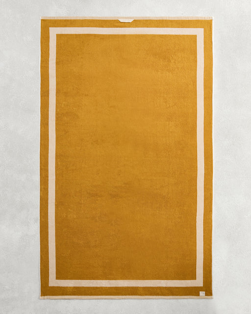The Bed Threads Crème & Turmeric Two-Toned 100% Cotton Terry Bath Sheet, featuring a golden-yellow and white design, is shown laid flat on a light gray textured surface.