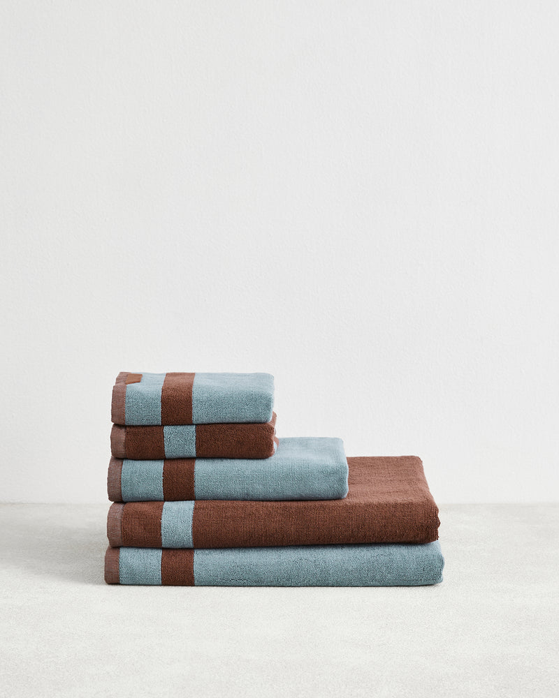 The Bed Threads Cacao & Lagoon Two-Toned 100% Cotton Terry Bath Sheet Bundle features four plush bath towels, neatly stacked from largest to smallest, in alternating blue and brown, displayed on a plain white background.