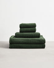 The Bed Threads Moss 100% Cotton Terry Bath Sheet Bundle, featuring bath sheets, towels, and hand towels in dark green, is neatly stacked on a light surface against a plain white background. - view