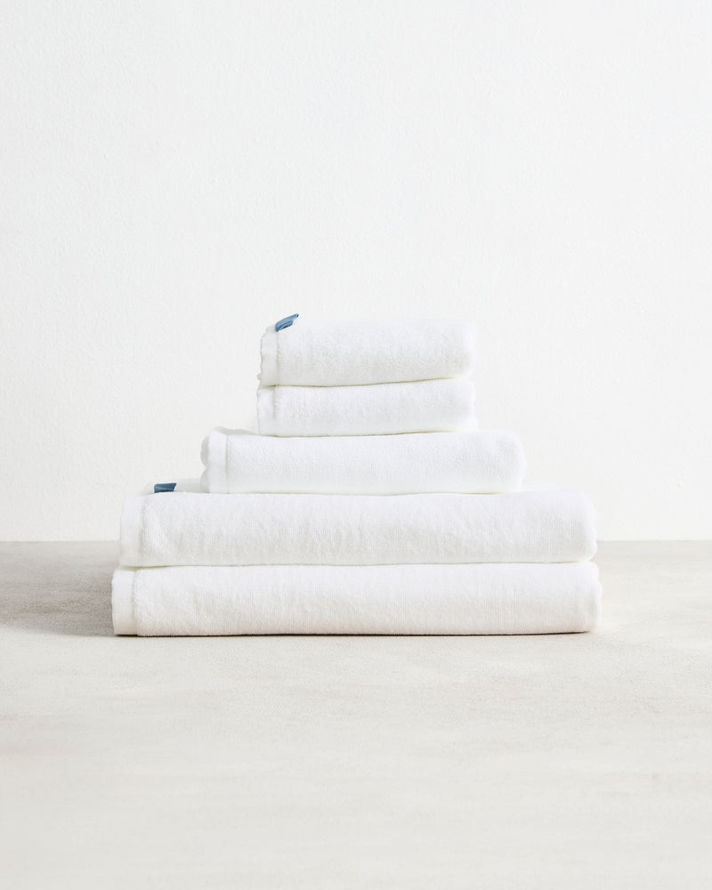 The Bed Threads Crisp White 100% Cotton Terry Bath Sheet Bundle features highly absorbent towels in various sizes, neatly stacked on a light surface against a plain white background.