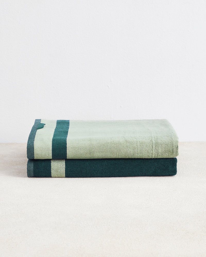 The Bed Threads Forest & Sage Two-Toned 100% Cotton Terry Bath Sheets set features two plush, quick-drying towels—one light green with a dark green stripe, one dark green with a light green stripe—stacked on a light surface against a white background.