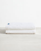 A pair of Crisp White 100% Cotton Terry Bath Sheets by Bed Threads, neatly folded with a blue tag, rests on a light textured surface against a plain white background. - view