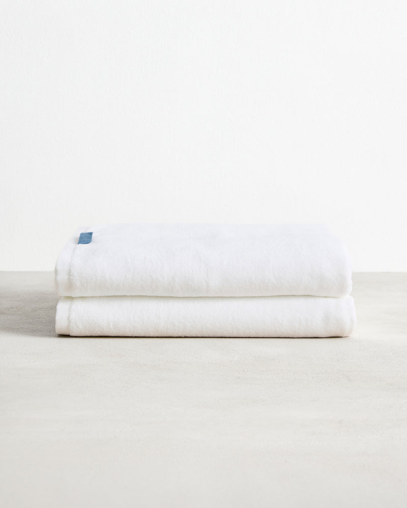A pair of Crisp White 100% Cotton Terry Bath Sheets by Bed Threads, neatly folded with a blue tag, rests on a light textured surface against a plain white background.