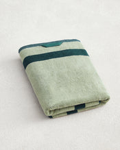 A Forest & Sage Two-Toned 100% Cotton Terry Bath Towel by Bed Threads, featuring light and dark green stripes and quick-drying cotton, is folded on a textured off-white surface. - view