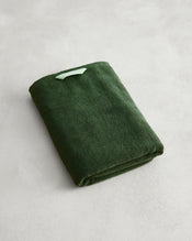 A folded Moss 100% Cotton Terry Bath Towel by Bed Threads in dark green rests on a light grey textured surface, with a small light green tag attached to its corner. - view