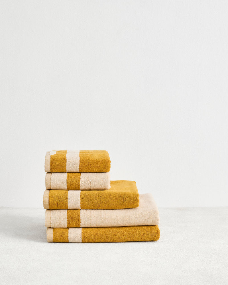 The Bed Threads Crème & Turmeric Two-Toned 100% Cotton Terry Bath Towel Bundle features four plush towels with a white stripe, neatly stacked on a light surface against a plain white wall.