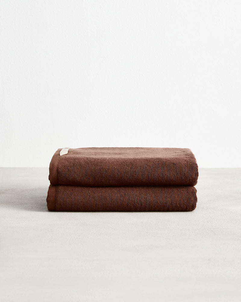 A set of two Bed Threads Espresso 100% Cotton Terry Bath Towels, neatly folded with a small beige tag, rests on a light gray surface against a white background.