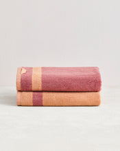 A set of two Bed Threads Terracotta & Pink Clay Two-Toned 100% Cotton Terry Bath Towels, one with a gold stripe and the other with a pink stripe, neatly folded on a light surface against a pale background. - view