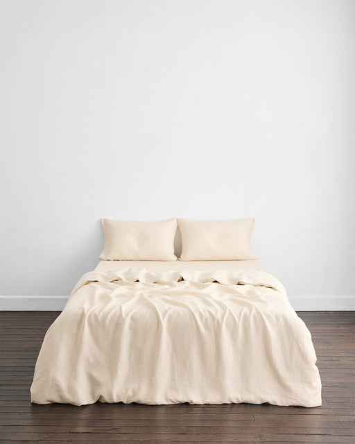 A minimalist room features a neatly made bed with the Bed Threads Crème 100% French Flax Linen Duvet Cover and two matching pillows, set against a plain white wall on a dark wooden floor.
