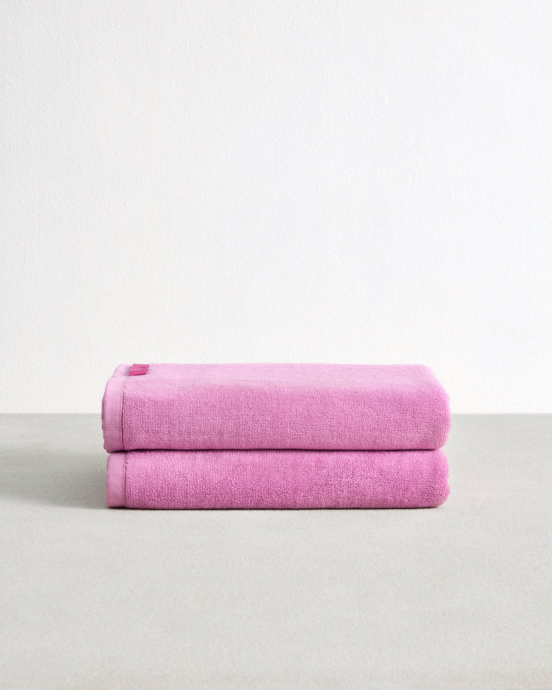 A set of two Bubblegum 100% Cotton Terry Bath Towels by Bed Threads, neatly folded and highly absorbent, are stacked on a light gray surface against a plain white background.