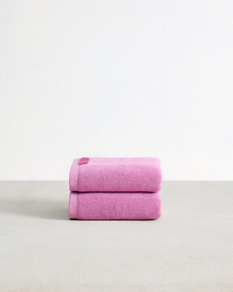 Two neatly folded Bubblegum 100% Cotton Terry Hand Towels by Bed Threads are stacked on a light gray surface with a plain white background. This highly absorbent set of two adds comfort and style to your daily routine.