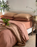 A neatly made bed features the Hazelnut & Terracotta 100% French Flax Linen Bedding Bundle from Bed Threads, beside a wooden stool with a potted plant. More greenery and natural light create a cozy, inviting, temperature-regulating atmosphere.