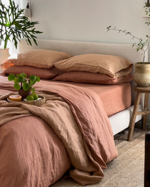 A neatly made bed features the Hazelnut & Terracotta 100% French Flax Linen Bedding Bundle from Bed Threads, beside a wooden stool with a potted plant. More greenery and natural light create a cozy, inviting, temperature-regulating atmosphere.