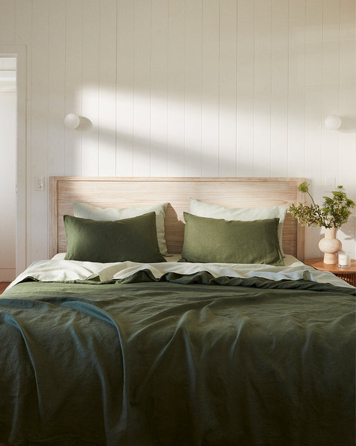 A bed dressed in Bed Threads’ Sage 100% French Flax Linen Sheet Set rests against a light wood-paneled wall, with a wooden nightstand holding a vase of greenery beside it, softly lit by natural sunlight.