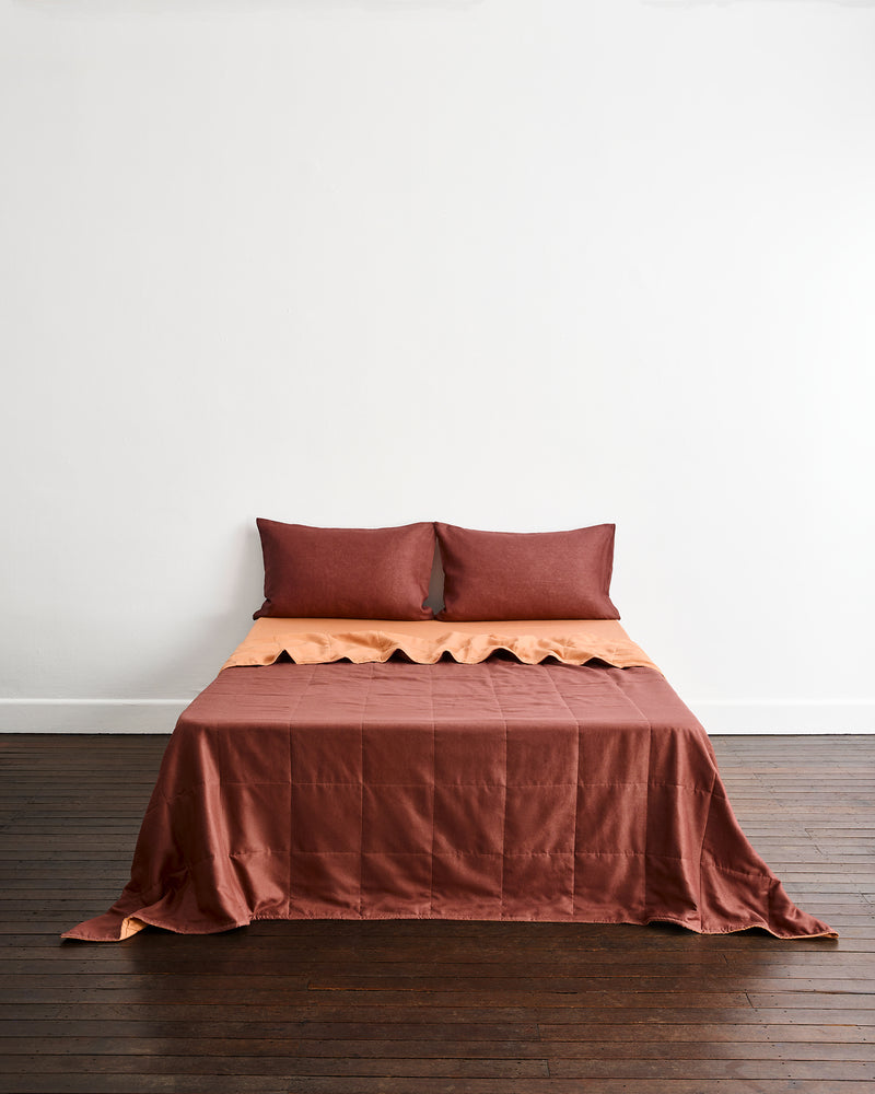 A bed dressed in a rust-brown quilt cover in Cacao & Hazelnut, with matching pillowcases in Cacao, set against a plain white wall and dark wood floor.
