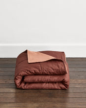 A folded rust-brown quilt cover in Cacao & Hazelnut with a hazelnut underside, displayed on a dark wooden floor against a plain white wall. - view
