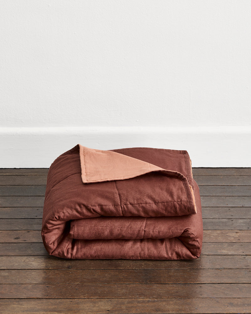 A folded rust-brown quilt cover in Cacao & Hazelnut with a hazelnut underside, displayed on a dark wooden floor against a plain white wall.